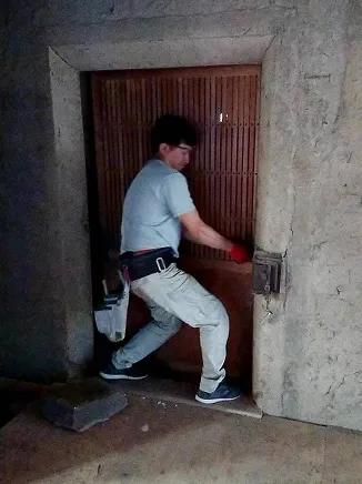 作業着の男性が赤い手袋で木製のドアを開けようとしている。