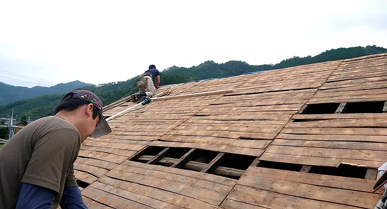 屋根の木製下地の上で屋根修理をする作業員二人。